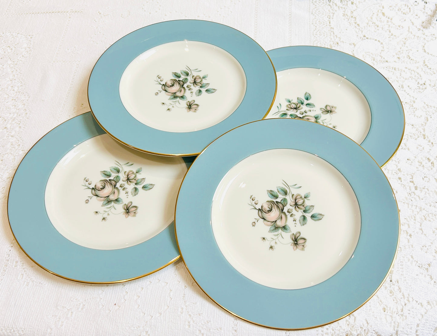 Four vintage Royal Doulton Rose Elegans TC1010 plates, each with a light blue border, gold trim, and floral centre, displayed on a lace tablecloth